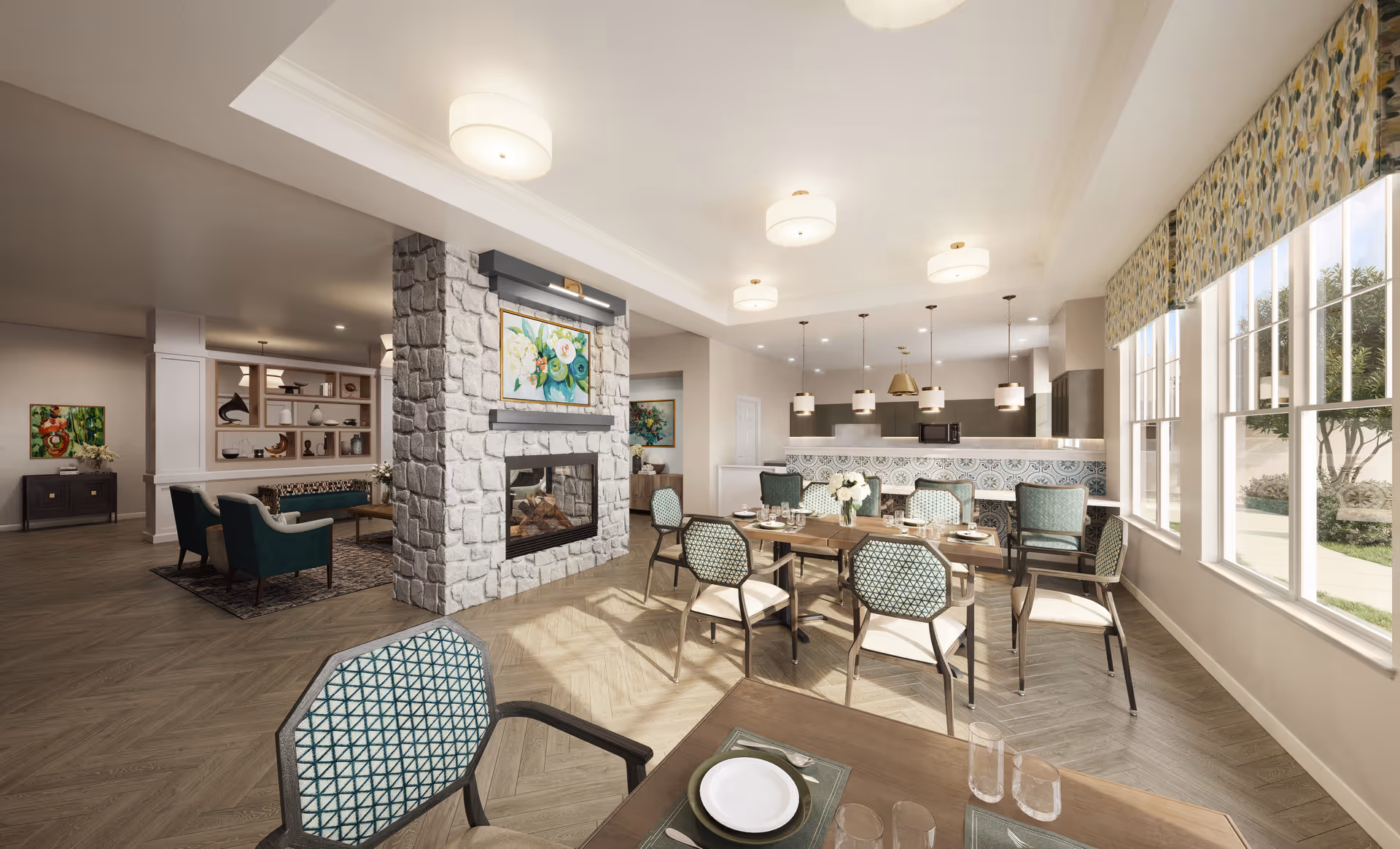 A bright and spacious dining area in an assisted living facility with multiple tables and chairs set for meals. The room features large windows with patterned valances letting in natural light, a stone fireplace with a colorful floral painting above it, and a kitchen area with pendant lighting and a decorative tile backsplash in the background. Adjacent to the dining area is a cozy seating area with armchairs and a rug.