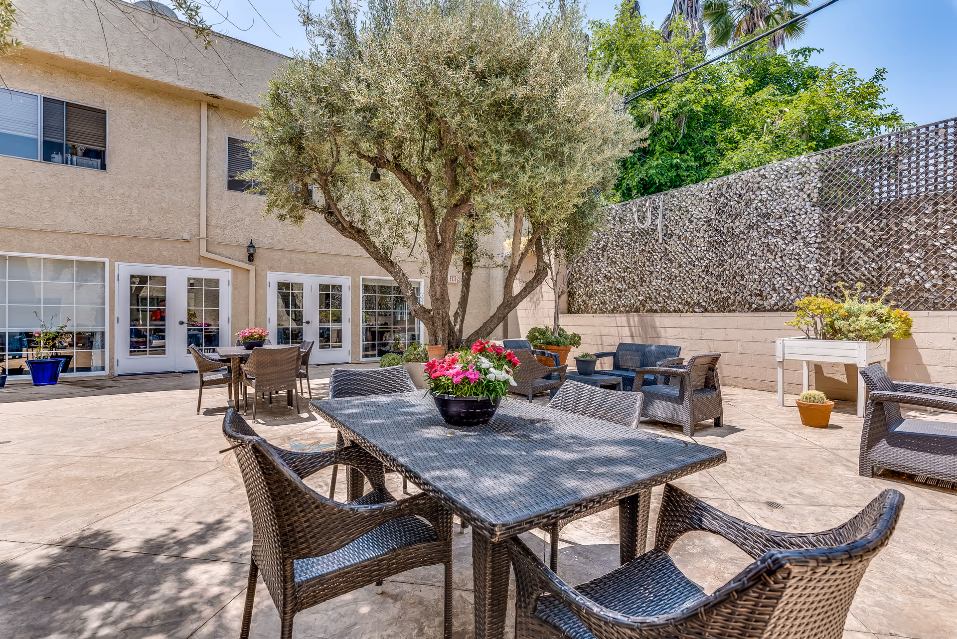 Outdoor patio area at Astoria Park Senior Living with multiple wicker tables and chairs arranged around potted plants and flowers. A large tree provides shade, and the patio is surrounded by a beige building and a privacy fence with greenery.