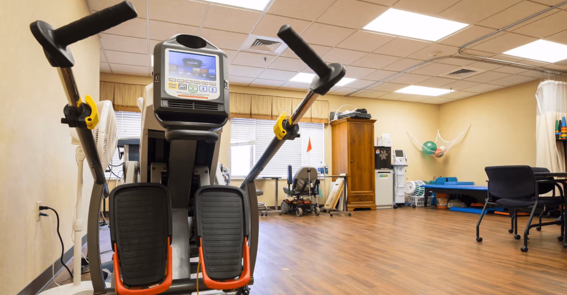 A spacious interior therapy/fitness room with an elliptical machine in the foreground and chairs, therapy equipment, and exercise balls along the walls.