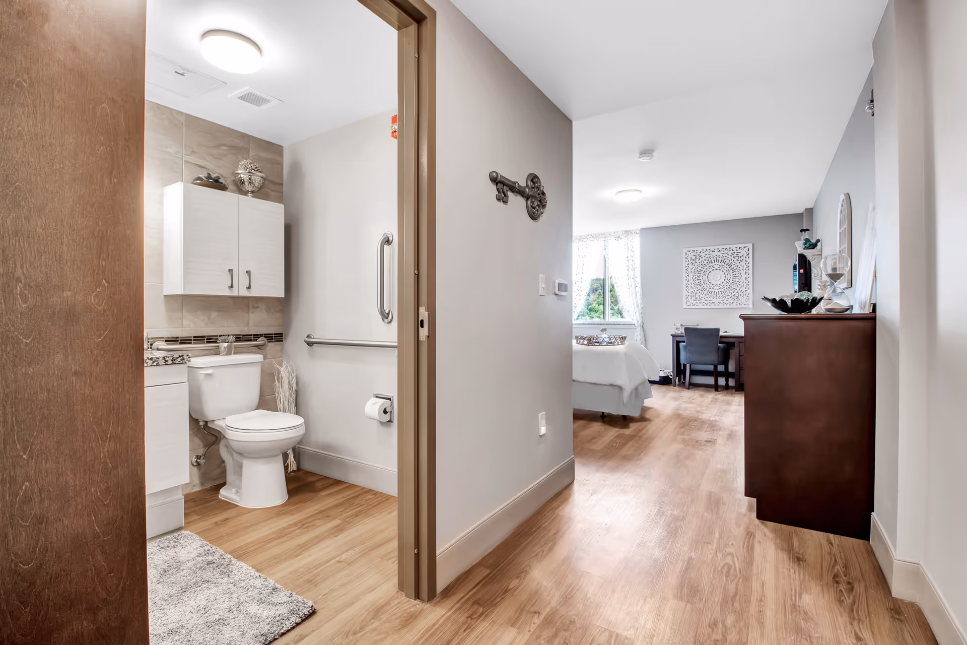 View of a hallway in an apartment showing a bathroom on the left with a toilet, white cabinet, and grab bars. The hallway leads to a bedroom area with a bed, desk, chair, and dresser, with natural light coming through a window with sheer curtains.