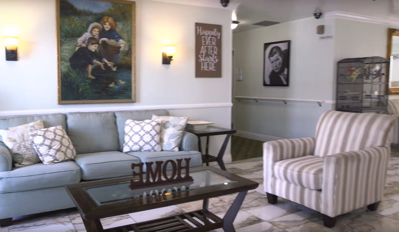 A cozy living room area with a light blue sofa adorned with patterned pillows, a striped armchair, and a glass coffee table with a decorative 'HOME' sign. The walls are decorated with a large painting of children by a pond, a framed black and white portrait, and a sign that reads 'Happily Ever After Starts Here'. The floor is tiled with a marble pattern, and a birdcage is visible in the background.
