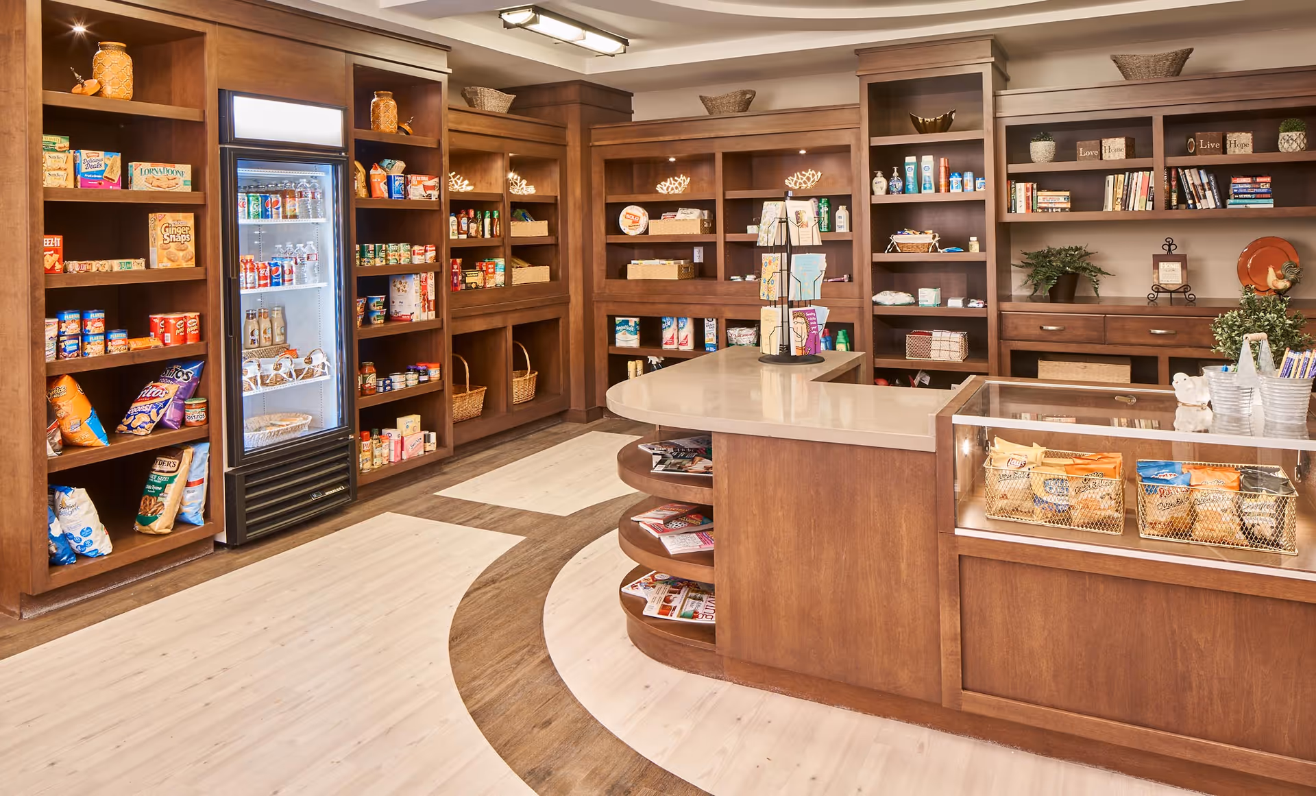 Interior view of a small convenience store or market area within a senior living facility, featuring wooden shelves stocked with snacks, canned goods, and beverages. A refrigerated display case holds drinks, and a wooden counter with baskets of packaged snacks is in the foreground. The space is well-lit with ceiling lights and has a light wood floor with a darker wood inlay design.