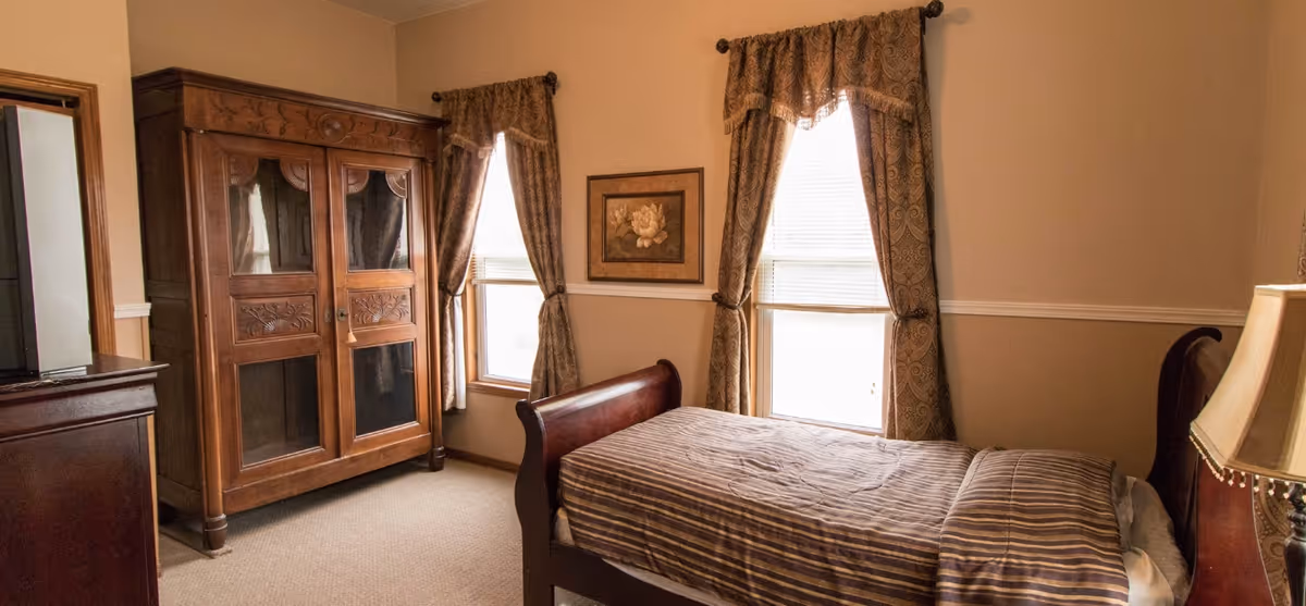 A bedroom with a wooden bed featuring a striped bedspread, two windows with patterned curtains, a wooden armoire with glass panels, a framed floral picture on the wall, and a lamp on a bedside table.