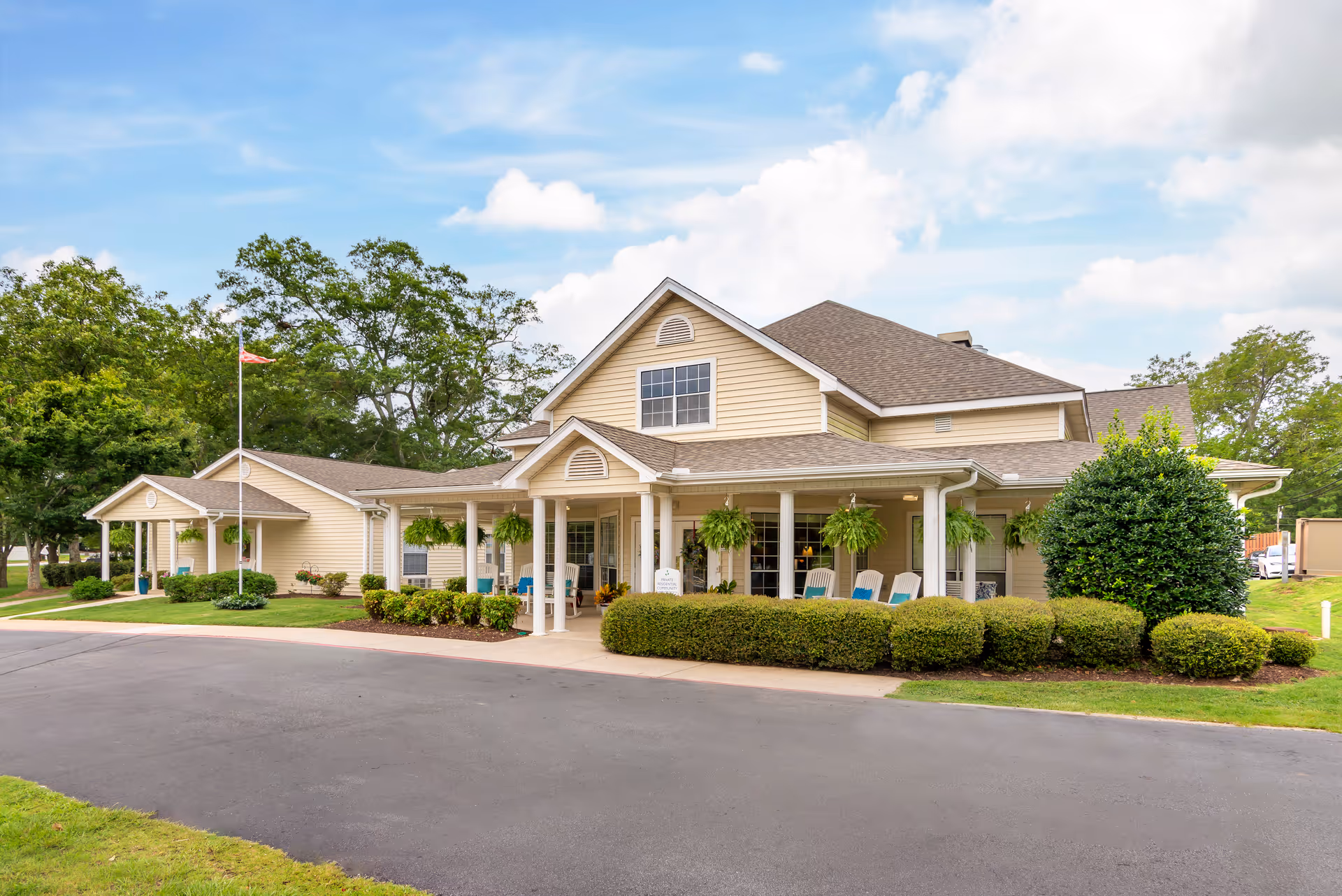 Exterior view of a single-story senior living facility building with beige siding, a covered porch with hanging plants, and several chairs. The building is surrounded by neatly trimmed bushes and trees under a partly cloudy sky.