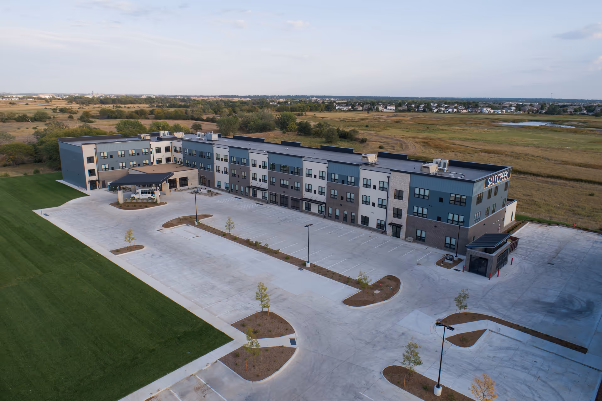 Aerial view of a three-story Salt Creek Senior Living building with a large empty parking lot and surrounding fields.