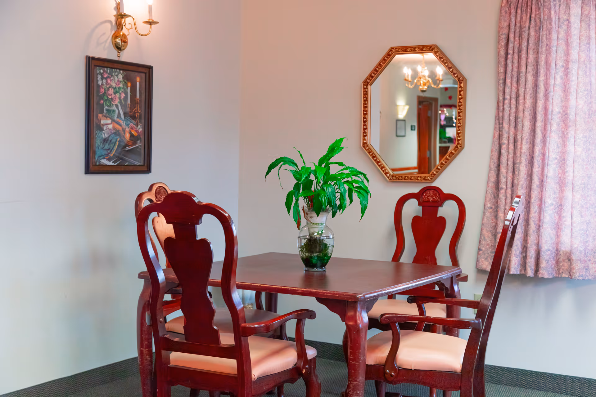 A small dining area with a square wooden table surrounded by four wooden chairs with cushioned seats. A green potted plant is placed in the center of the table. On the wall behind the table, there is an octagonal mirror and a framed painting of flowers and a violin. A wall sconce light fixture is mounted above the painting, and a window with floral curtains is visible on the right side.