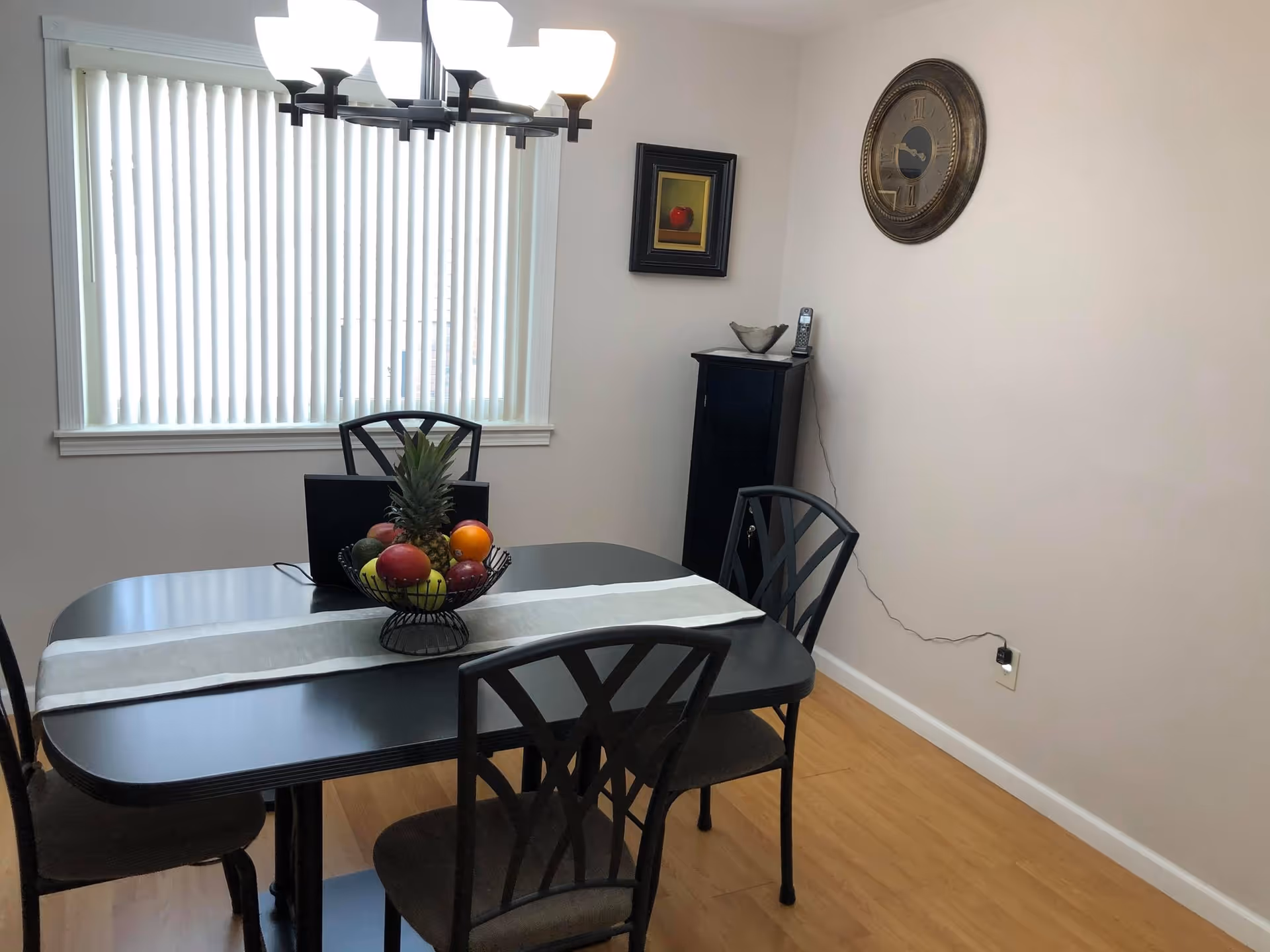 Dining room with a black table and four chairs, a fruit bowl centerpiece, vertical blinds over a window, and a wall clock.