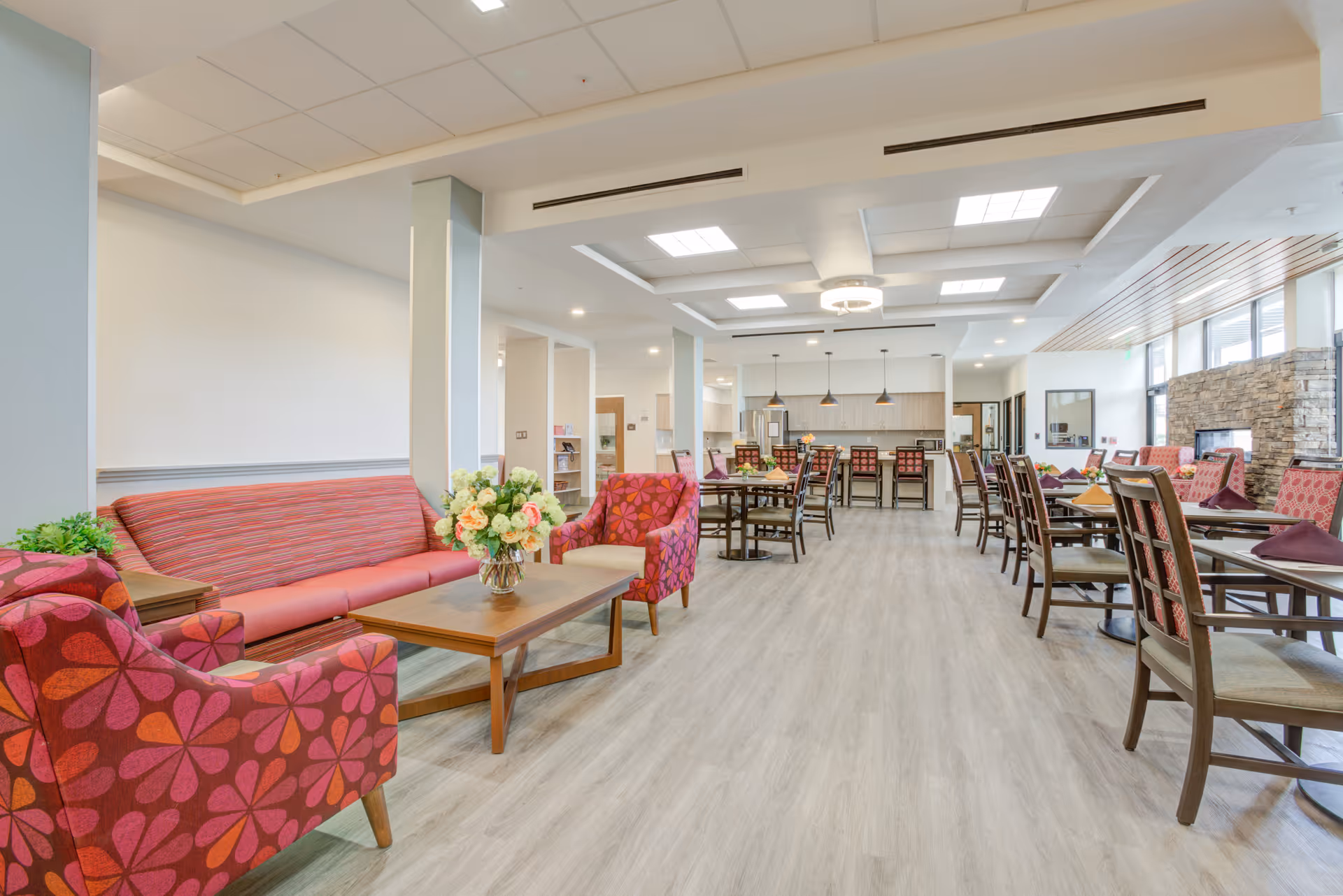 A spacious and well-lit senior living common area featuring a seating area with a red patterned sofa and armchairs around a wooden coffee table with a flower vase. In the background, there are multiple dining tables with chairs and a kitchen area with pendant lights and bar stools. The room has large windows, a stone fireplace, and light wood flooring.