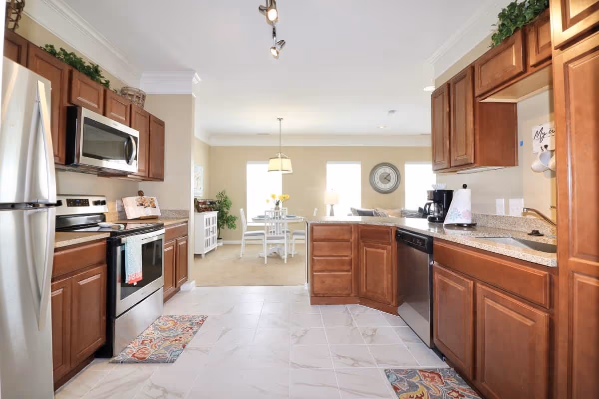 Bright kitchen with wooden cabinets, stainless steel refrigerator, oven, microwave, and dishwasher. The kitchen has a tiled floor with colorful rugs and granite countertops. In the background, there is a dining area with a white table and chairs, a hanging light fixture, and a wall clock.