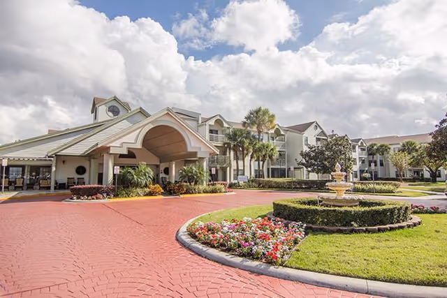Exterior view of Cascade Heights senior living facility showing a large covered entrance with a circular driveway, landscaped garden with colorful flowers, a fountain, palm trees, and a multi-story building under a partly cloudy sky.