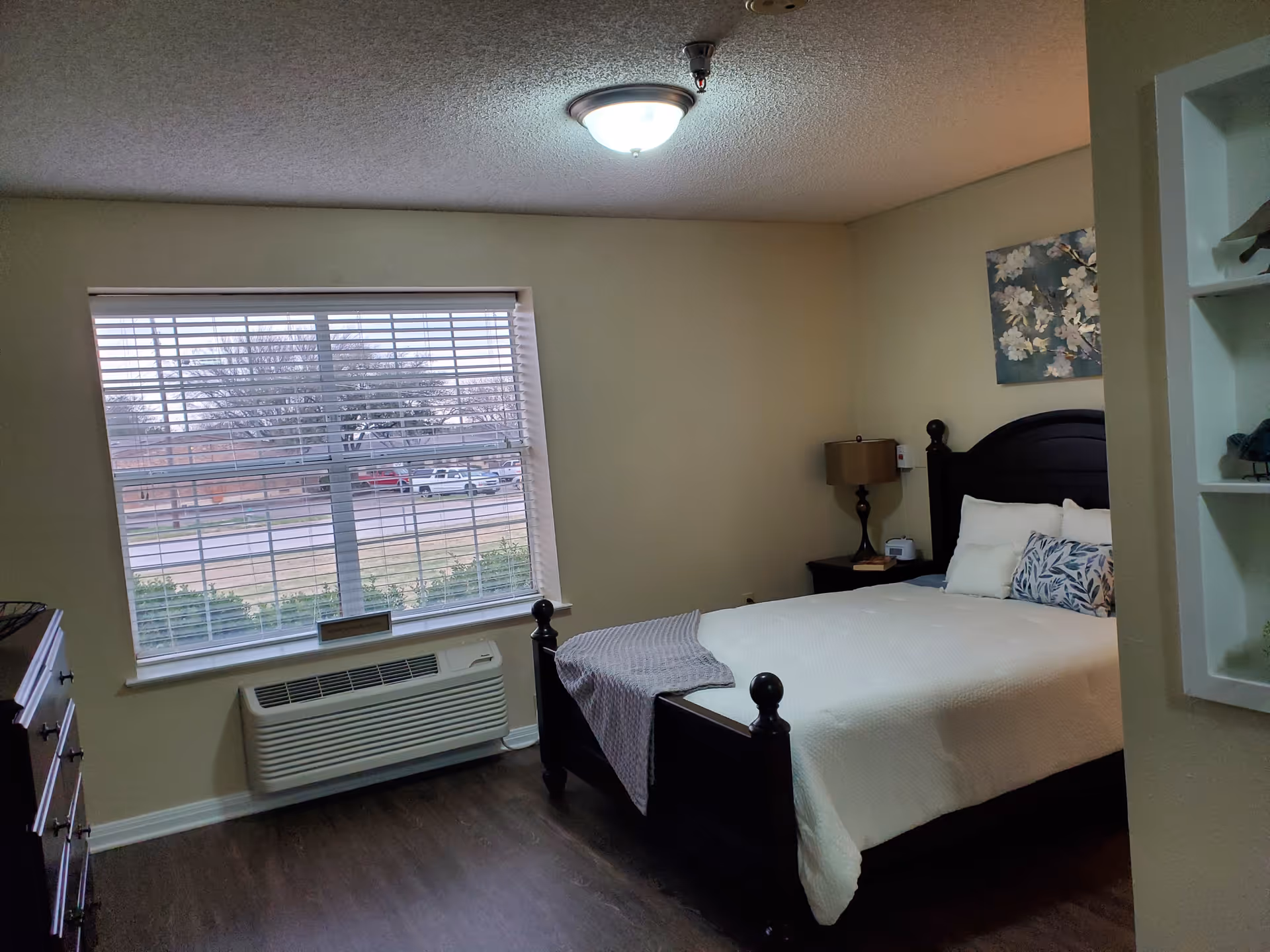 A furnished bedroom with a bed, bedside table and lamp, large window with blinds, and an air conditioning unit below the window.