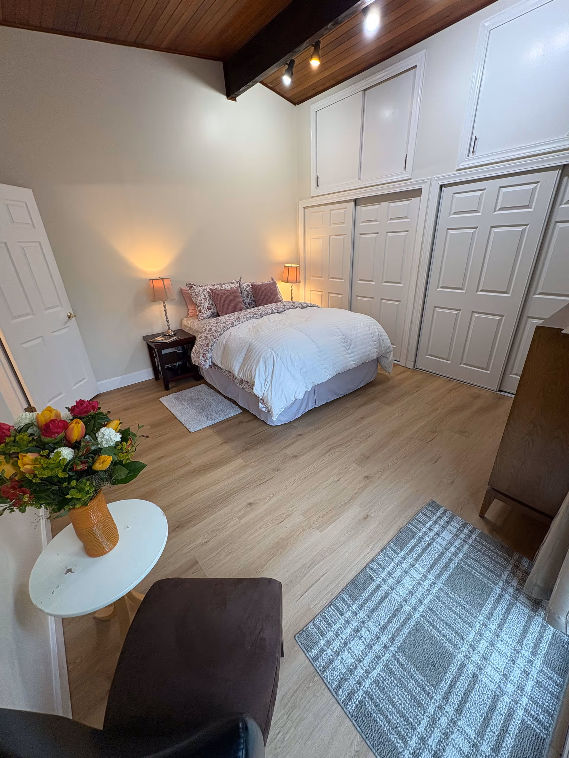 A cozy bedroom with a bed featuring white and floral bedding, two bedside tables with lamps, a wooden dresser, a plaid rug on the wooden floor, a small round table with a vase of colorful flowers, and a chair. The room has high ceilings with wooden beams and multiple closet doors.