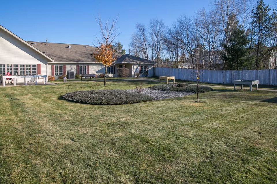 A well-maintained outdoor garden area at Cranberry Court Assisted Living with a large grassy lawn, a few small trees, and a central landscaped section with shrubs and rocks. The building is visible in the background with beige siding and red shutters, under a clear blue sky.