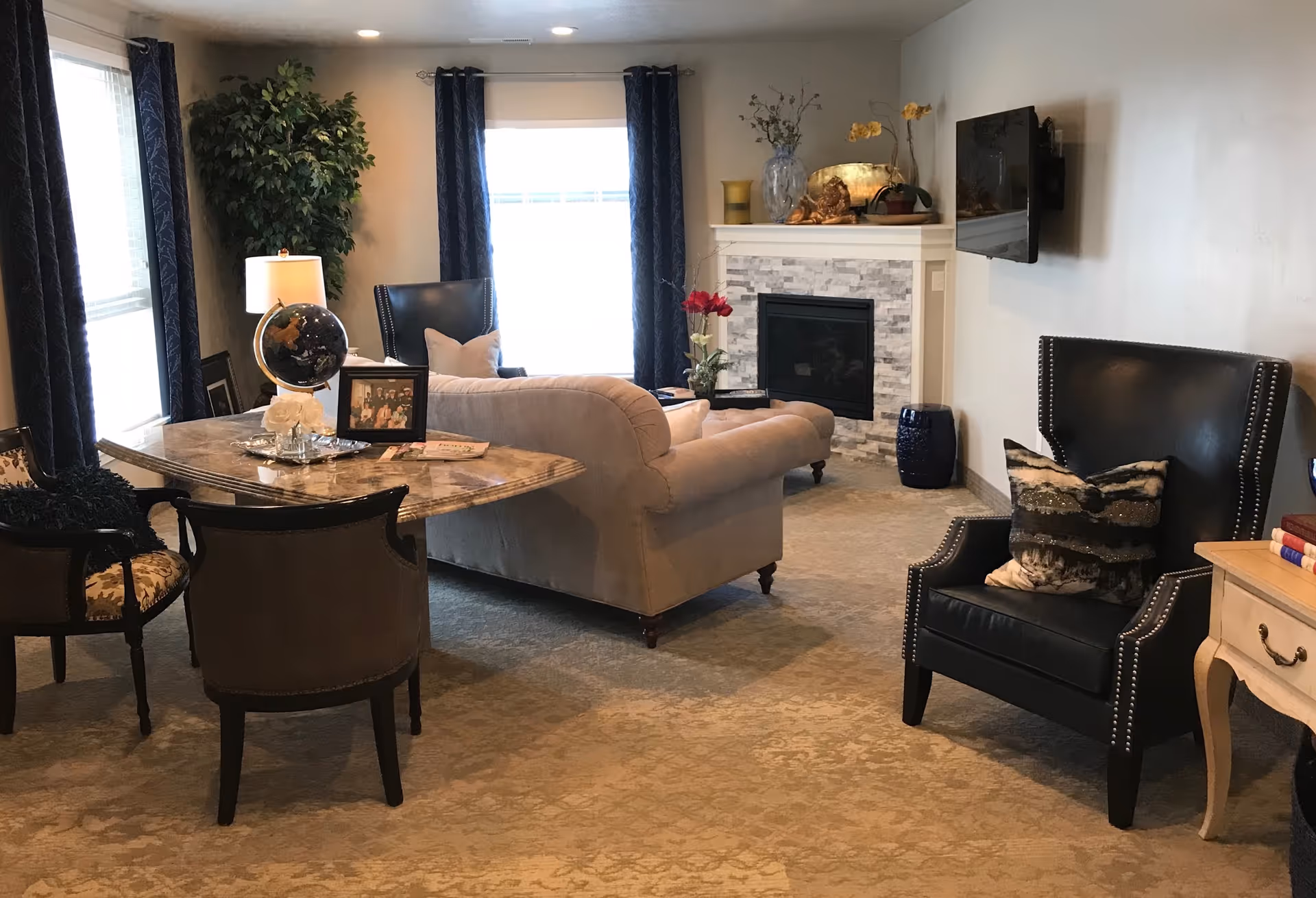 A cozy living room with a beige sofa, a black leather armchair with a decorative pillow, a marble-top table with chairs, a lamp, a globe, framed photos, and a fireplace with decorative items on the mantel. The room has carpeted flooring and windows with dark curtains.