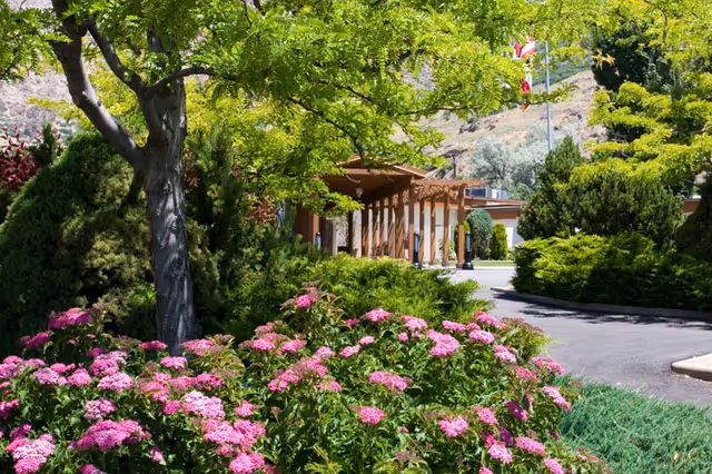 A lush garden area with vibrant pink flowers and green shrubs in the foreground, a tree with bright green leaves, and a pathway leading to a building entrance with wooden pergola structures. Hills and a flagpole with an American flag are visible in the background under a clear sky.