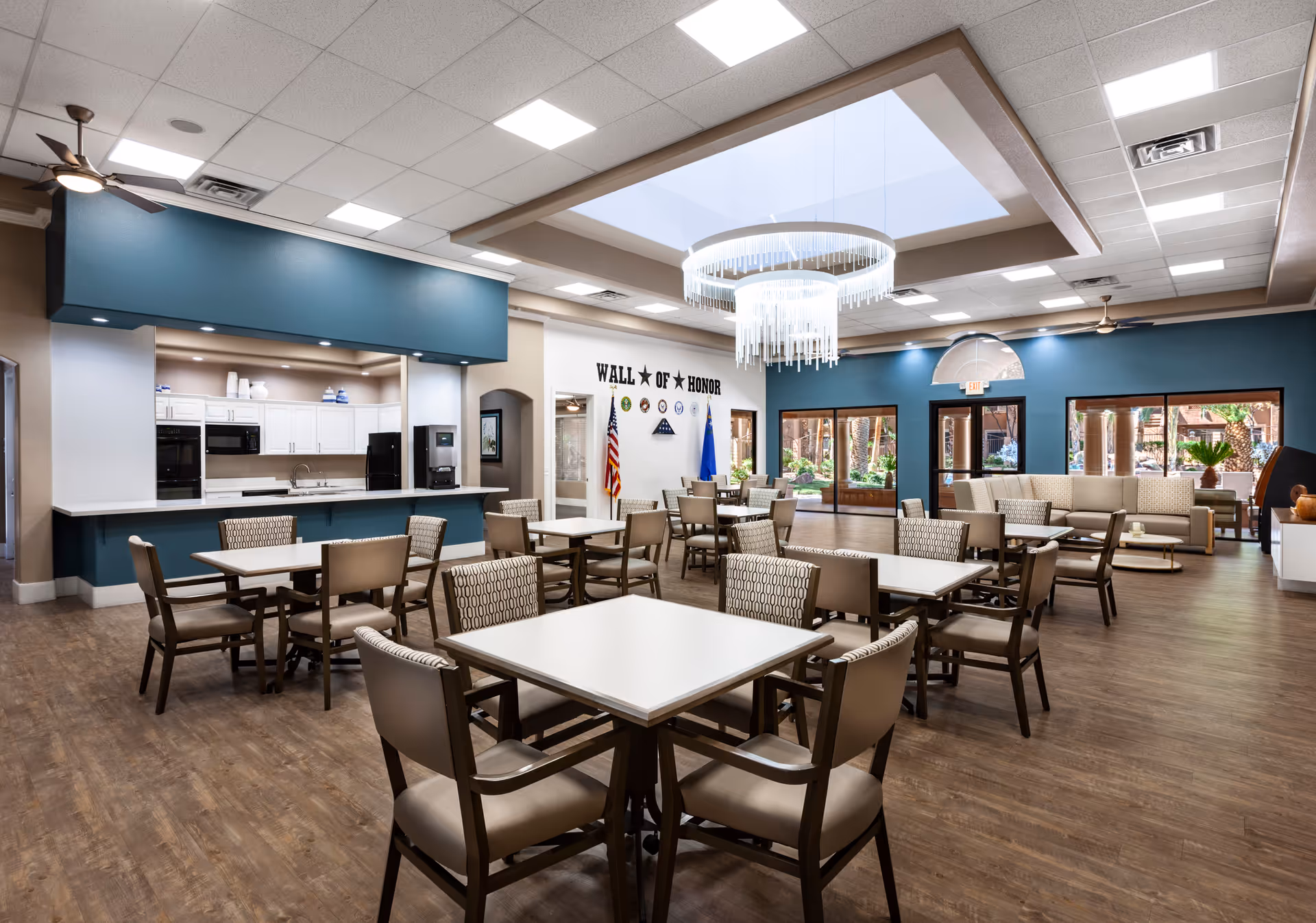 A spacious dining area in a senior living facility with multiple tables and chairs arranged neatly. The room features a modern kitchen area with white cabinets and black appliances on the left side. A large chandelier hangs from a skylight in the ceiling. There is a 'Wall of Honor' with flags and military insignia on the far wall. Large windows and glass doors provide a view of an outdoor patio area with greenery.