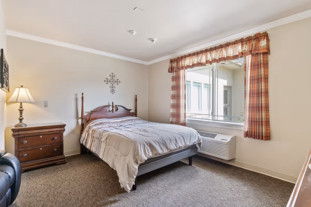 A simple bedroom with a wooden bed frame and a beige comforter. There is a wooden nightstand with a lamp on the left side of the bed. A window with plaid curtains lets in natural light, and a heating/cooling unit is installed below the window. The walls are light-colored, and the floor is carpeted.