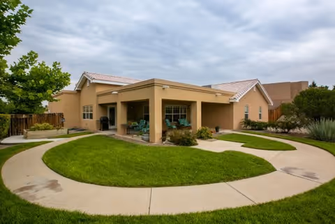 Single-story beige building with a covered patio area furnished with chairs, surrounded by a curved concrete walkway and well-maintained green lawn under a cloudy sky.