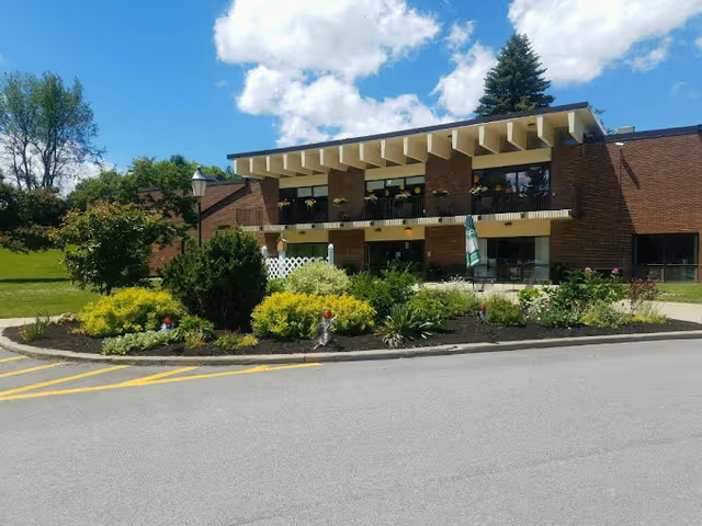 Exterior view of a brick residential care center building with a landscaped garden in front, featuring various shrubs and flowers. The building has a balcony with flower pots and large windows, under a partly cloudy blue sky.