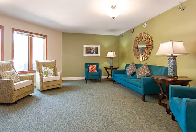 A cozy living room with green walls and carpeted floor featuring a teal sofa with patterned pillows, two teal armchairs, two beige armchairs with decorative pillows, two wooden side tables with lamps, a round decorative mirror on the wall, and a framed picture above one armchair. There are two large windows letting in natural light.