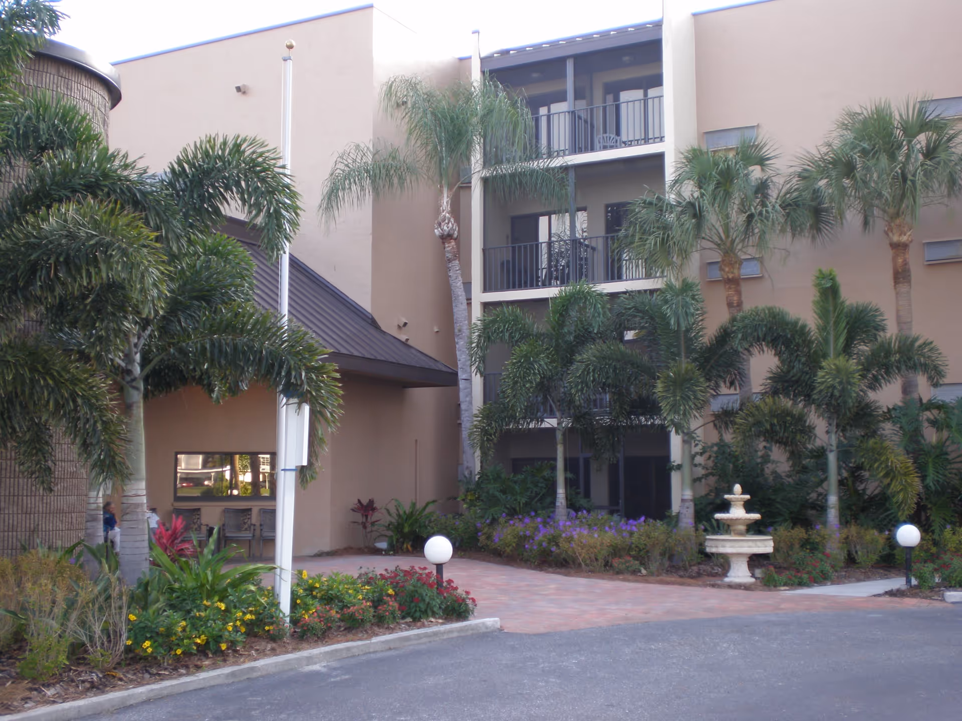 Exterior view of a multi-story building with balconies, surrounded by palm trees and landscaped flower beds. There is a small fountain in the garden area and a paved walkway leading to the entrance.