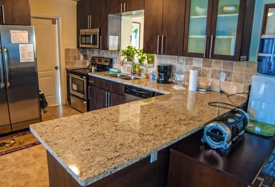 Modern kitchen with a large granite island, dark wood cabinets, stainless steel refrigerator and stove, and a sink with countertop appliances.