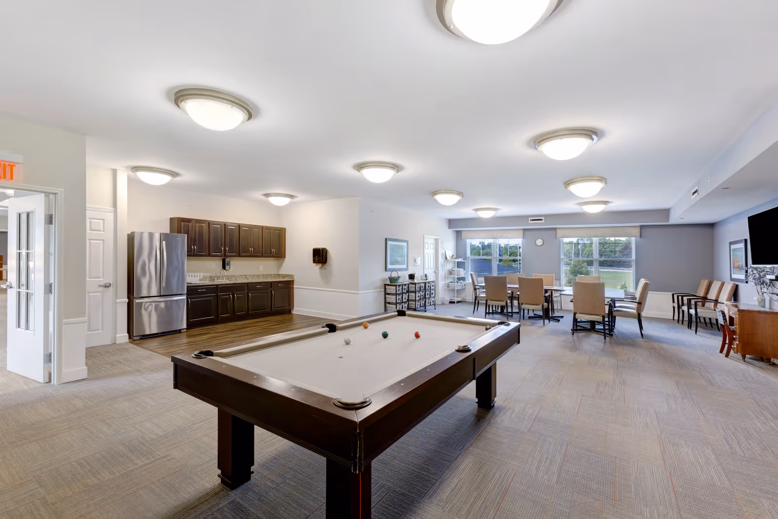 A spacious common room with a pool table in the foreground, a kitchenette with dark cabinets and a stainless steel refrigerator on the left, and a dining area with tables and chairs near large windows letting in natural light.
