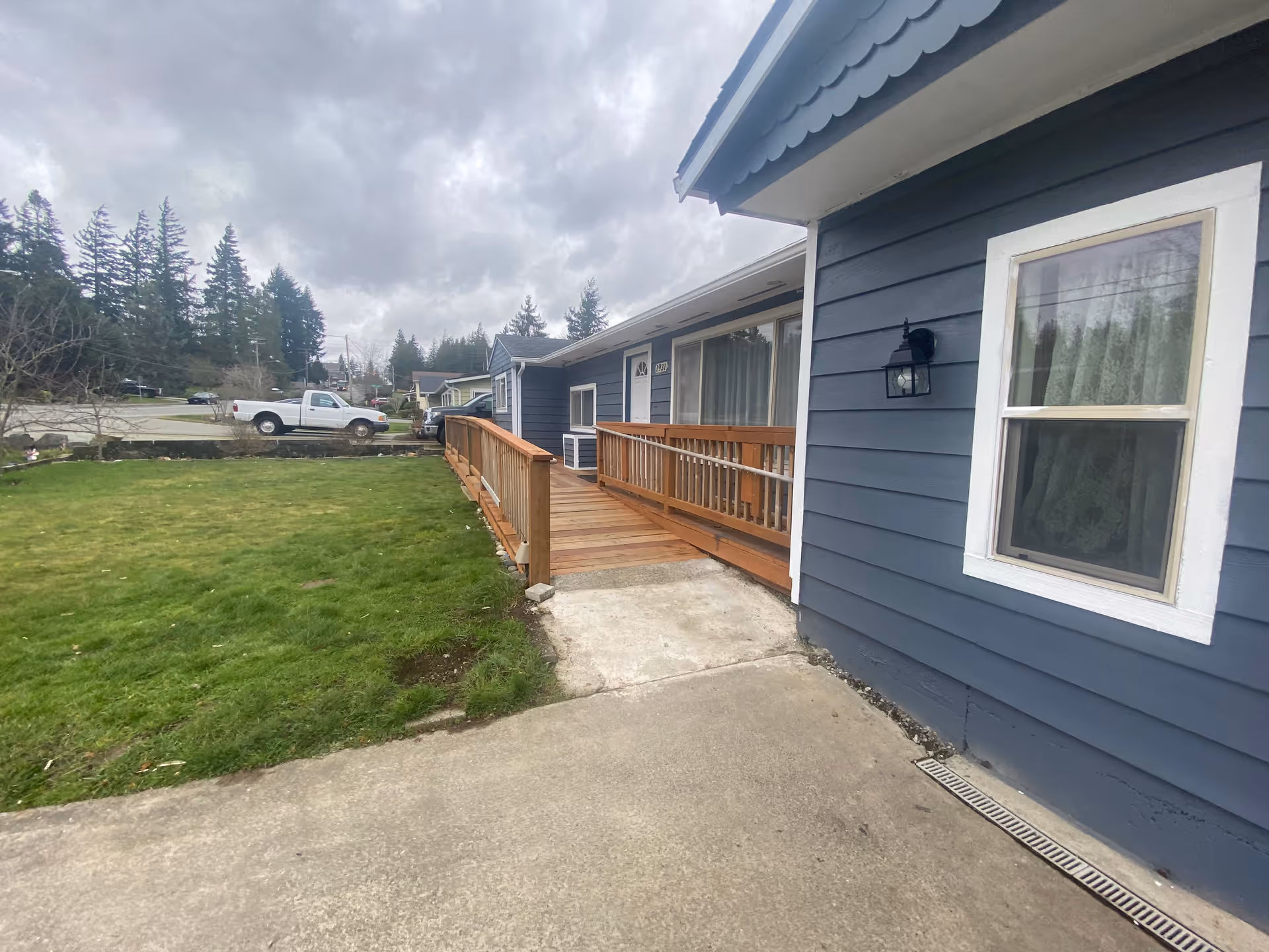 Front exterior of a blue single-story building with a wooden ramp, lawn, and driveway under a cloudy sky.