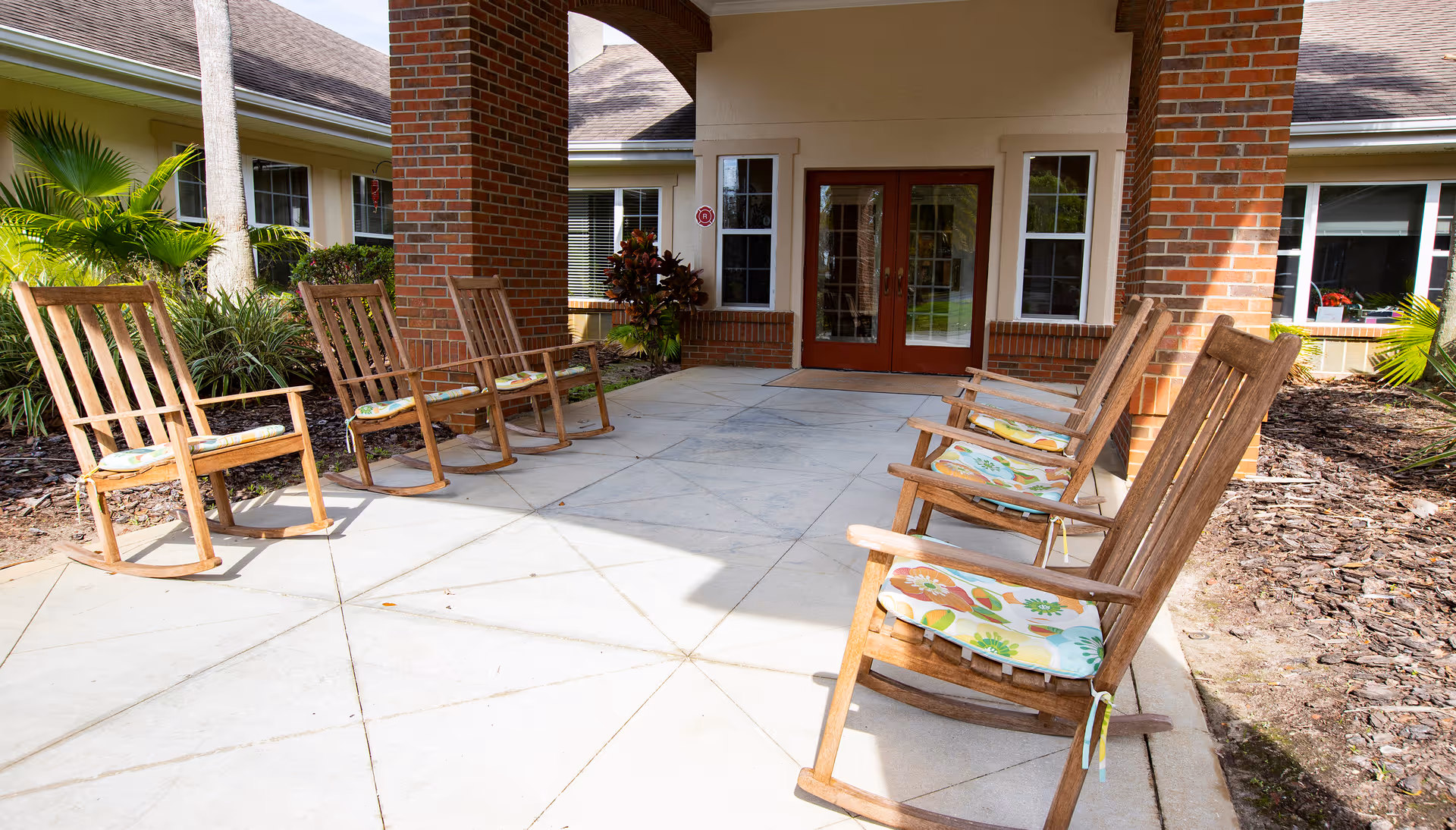 Outdoor covered patio area with six wooden rocking chairs arranged in two rows facing each other, each with a floral cushion. The patio is supported by brick columns and leads to a building entrance with double glass doors and windows on either side. Surrounding the patio are landscaped plants and mulch beds.