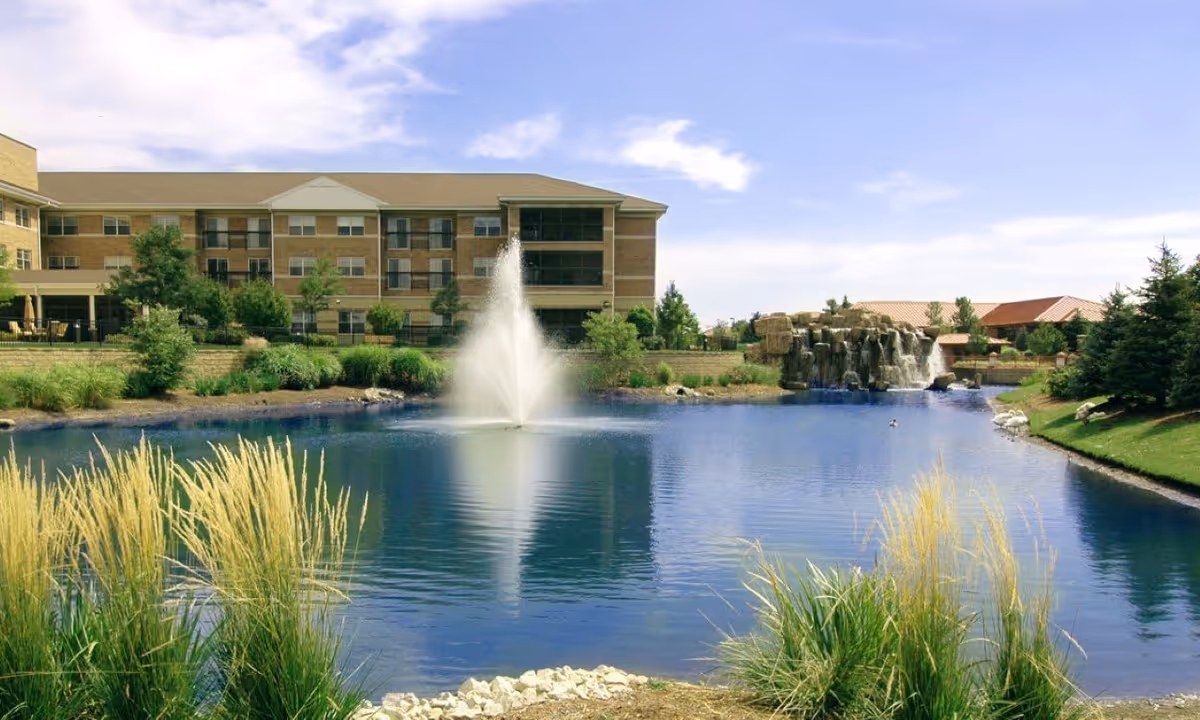 A serene outdoor scene at Alden of Waterford featuring a large pond with a central water fountain and a rock waterfall on the far side. The pond is surrounded by green grass, ornamental grasses, trees, and shrubs. In the background, there is a multi-story brick building under a partly cloudy blue sky.