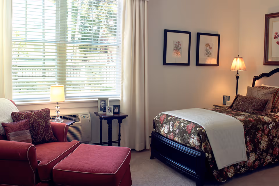 A cozy bedroom with a floral-patterned bedspread on a dark wooden bed frame, a red armchair with matching ottoman and decorative pillows, a small side table with framed photos and a lamp, and large windows with white blinds letting in natural light.