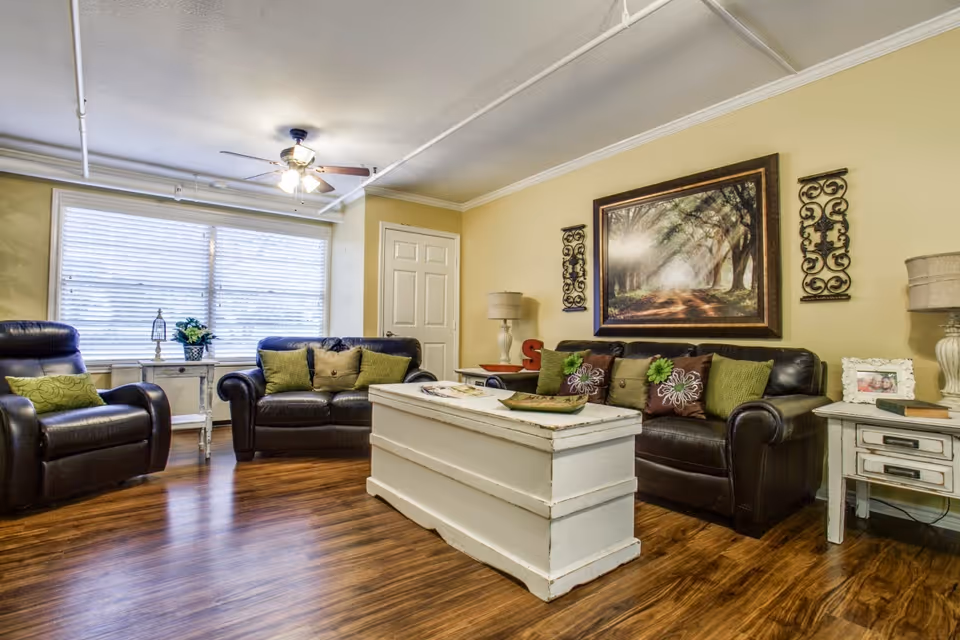 A cozy living room with two dark leather sofas adorned with green and brown decorative pillows. A white wooden chest serves as a coffee table in the center. The walls are painted a soft yellow and decorated with a large framed picture of a tree-lined path and two ornamental wall hangings. There are two side tables with lamps and decorative items, and a large window with blinds letting in natural light. The floor is a polished wood finish, and a ceiling fan with lights is mounted above.