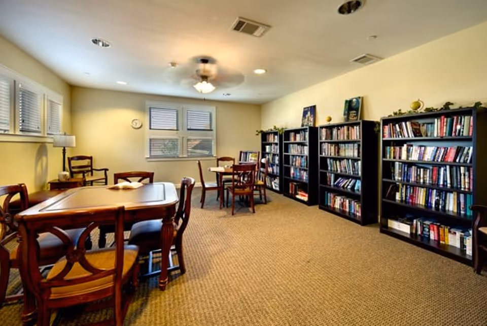 A cozy reading room with several wooden tables and chairs, bookshelves filled with books along one wall, windows with blinds, a ceiling fan with light, and a beige carpeted floor.