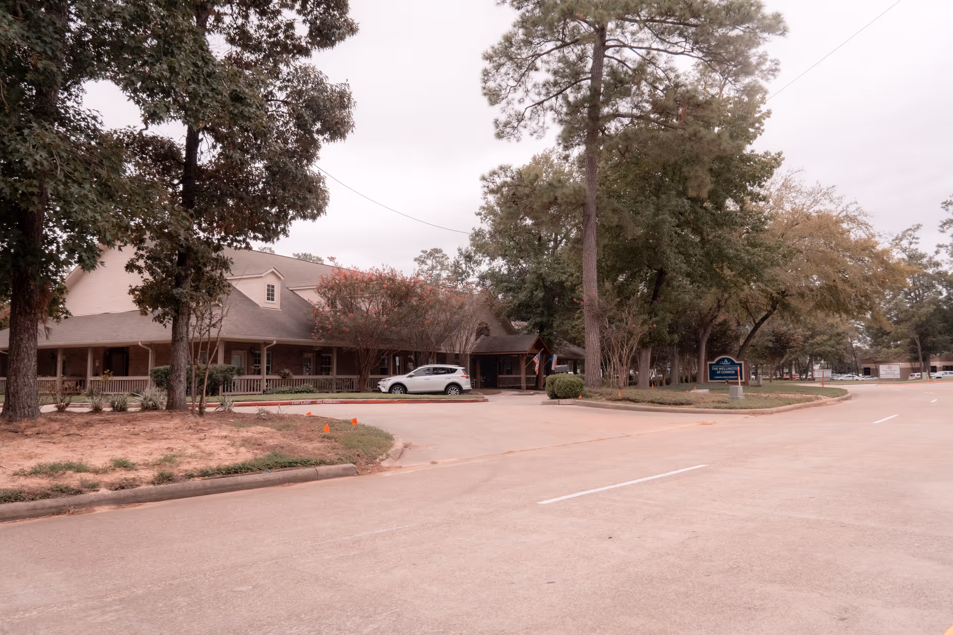 Exterior view of The Wellington at Conroe senior living facility showing a large building surrounded by trees and a parking area with a white SUV parked near the entrance. There is a sign for the facility on the right side near the road.
