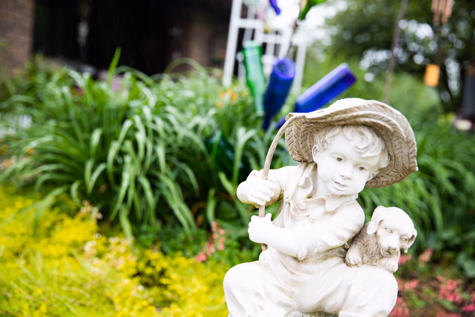 A white statue of a young boy wearing a hat, holding a stick, with a small dog sitting on his shoulder, set in a garden with green plants and colorful flowers in the background.