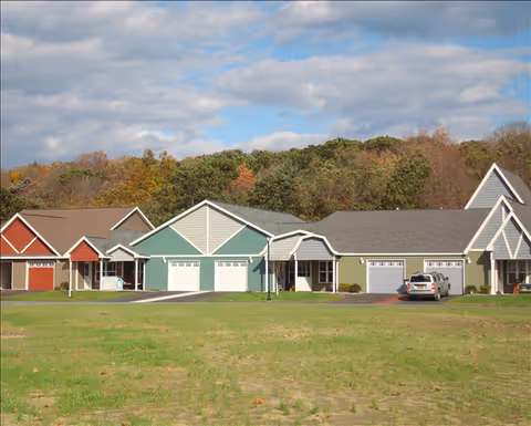Row of colorful single-story residential units with garages facing a grassy lawn and trees under a partly cloudy sky.