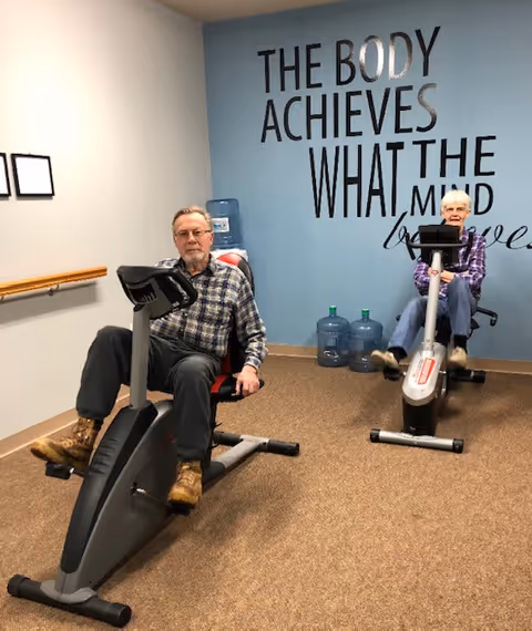 Two elderly individuals exercising on stationary recumbent bikes in a small fitness room with a blue wall that has motivational text reading 'THE BODY ACHIEVES WHAT THE MIND believes'. There are water jugs placed against the wall and a wooden handrail on the adjacent wall.