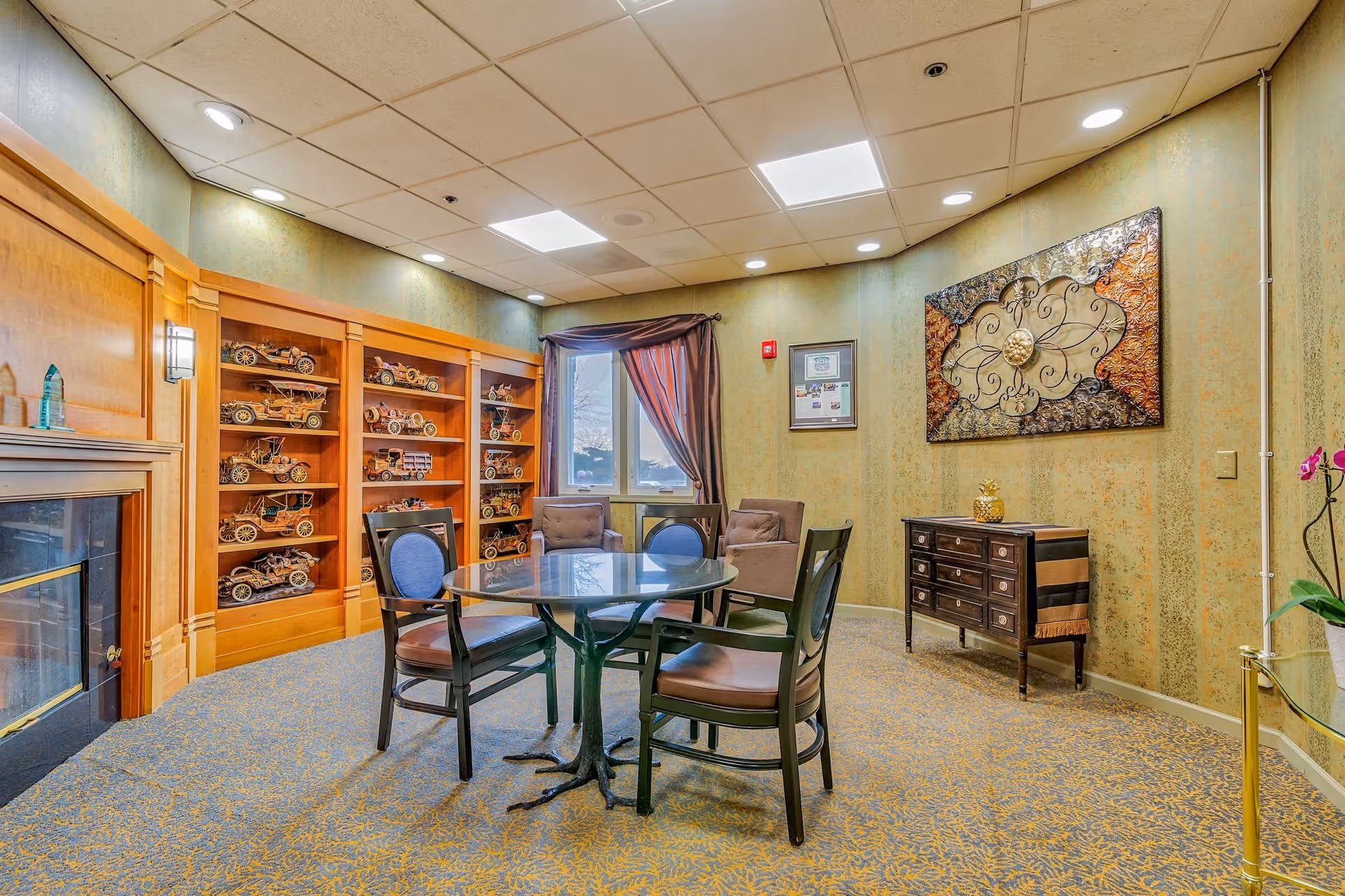 A cozy interior room with a round glass table surrounded by four chairs. The room features a wooden fireplace on the left, built-in wooden shelves filled with model cars, a window with brown curtains, a small chest of drawers with a decorative item on top, and a large decorative wall art piece. The carpet has a yellow and blue pattern, and the ceiling has recessed lighting.