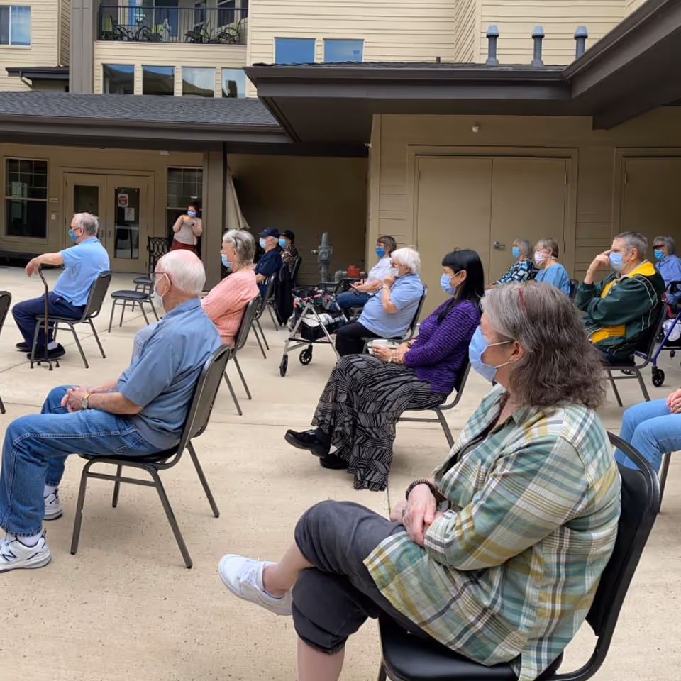 A group of elderly people sitting on chairs spaced apart in an outdoor courtyard area of a senior living facility. All individuals are wearing face masks and appear to be attentively listening or watching something. The background shows the exterior walls and windows of the building.