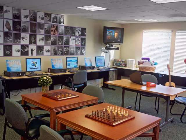 A senior living facility activity room with several tables set up for board games including chess and checkers. There are multiple computers along one wall, a wall-mounted television, and a window letting in natural light. The walls feature a display of black and white handprint photos. Chairs are arranged around the tables and computers.