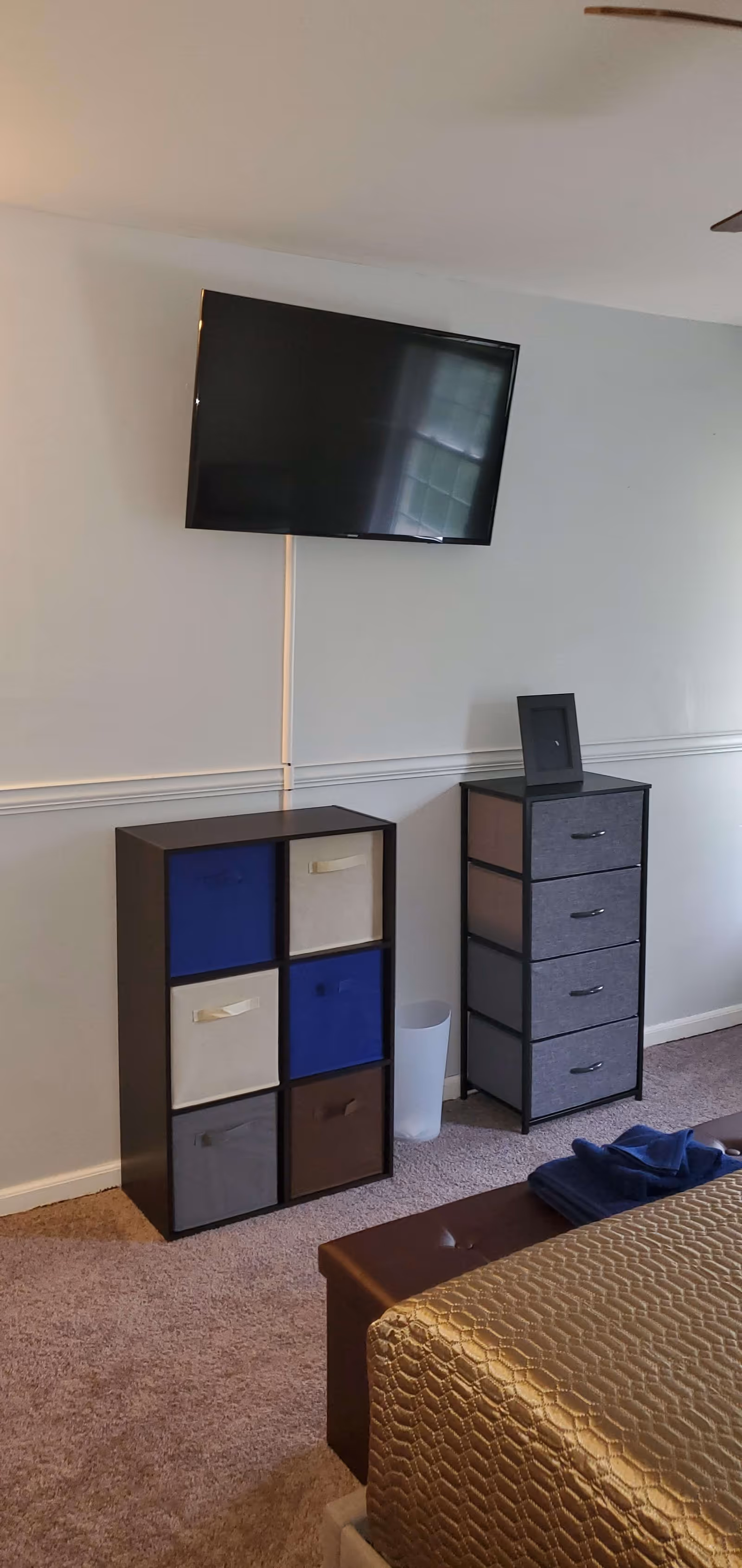 A bedroom corner with a wall-mounted flat-screen TV above a storage unit with six fabric drawers in blue, beige, gray, and brown. Next to it is a taller storage unit with four gray fabric drawers and a small picture frame on top. The floor is carpeted, and part of a bed with a gold textured bedspread and folded blue towels is visible in the foreground.