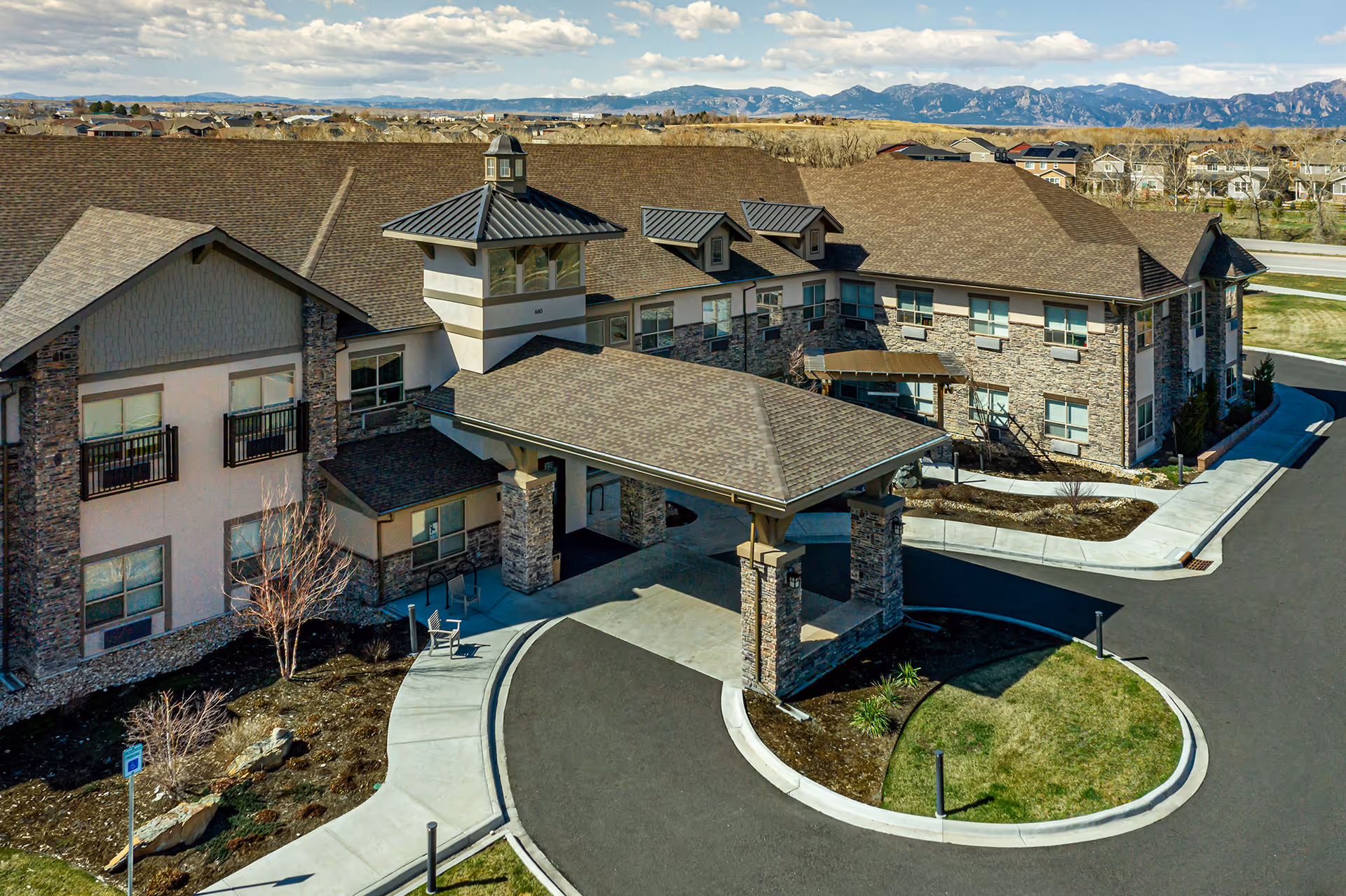 Exterior view of The Peaks at Old Laramie Trail senior living facility showing a large building with stone and beige siding, multiple windows, and a covered entrance with stone pillars. The driveway curves around a small grassy island, and mountains are visible in the background under a partly cloudy sky.