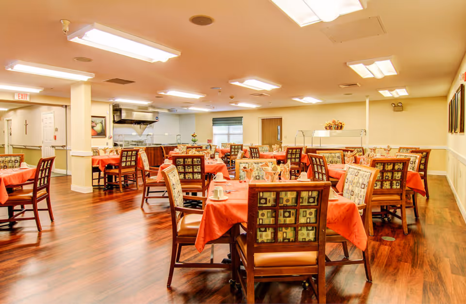 A dining room in a senior living facility with multiple tables covered in orange tablecloths. Each table is set with cups, napkins, and utensils. The chairs have patterned upholstery, and the room has wooden flooring and bright overhead lighting. There is a serving area with a food warmer in the background and a window with blinds.