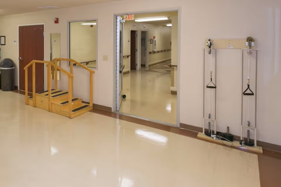 An indoor hallway area in a healthcare facility with a small wooden staircase with handrails, a closed wooden door, a trash can, and exercise equipment mounted on the wall. The hallway has beige walls and a shiny, light-colored floor, with an open doorway leading to another corridor.