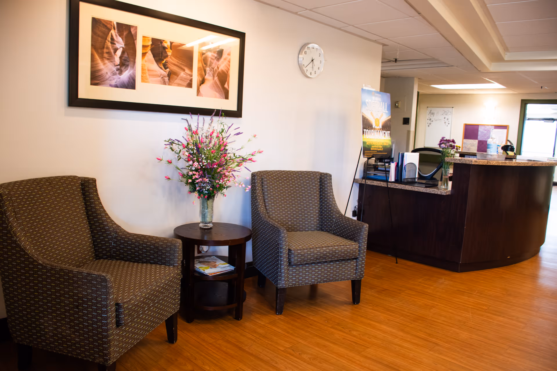 Reception/waiting area with two patterned armchairs flanking a small table holding a vase of flowers, wall art, and a reception desk in the background.