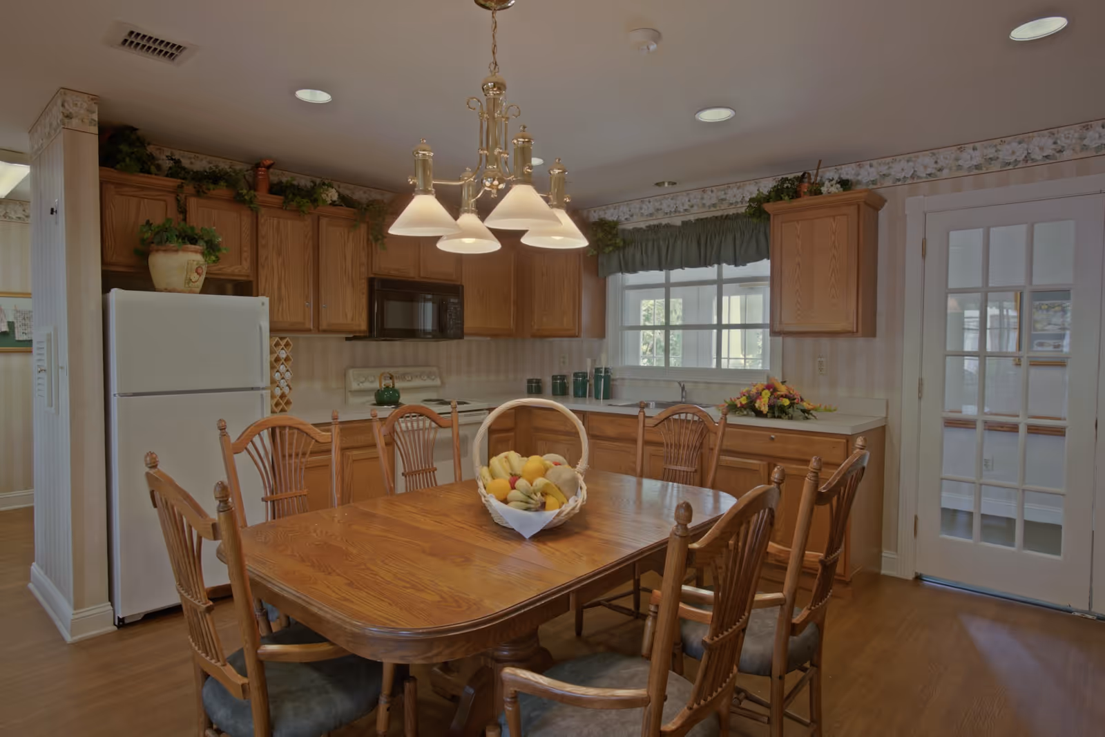 A kitchen and dining area with wooden cabinets, a white refrigerator, a microwave, and a stove. A wooden dining table with six chairs is centered in the room, with a basket of fruit on the table. There is a window with a green valance above the sink and a glass-paneled door to the right.
