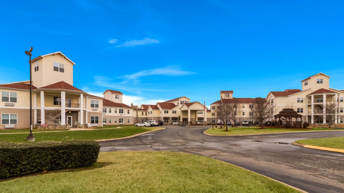 Exterior view of a large senior living facility named Fieldstone Place with multiple connected buildings featuring beige siding and red roofs, surrounded by green lawns, a paved driveway, and a clear blue sky.