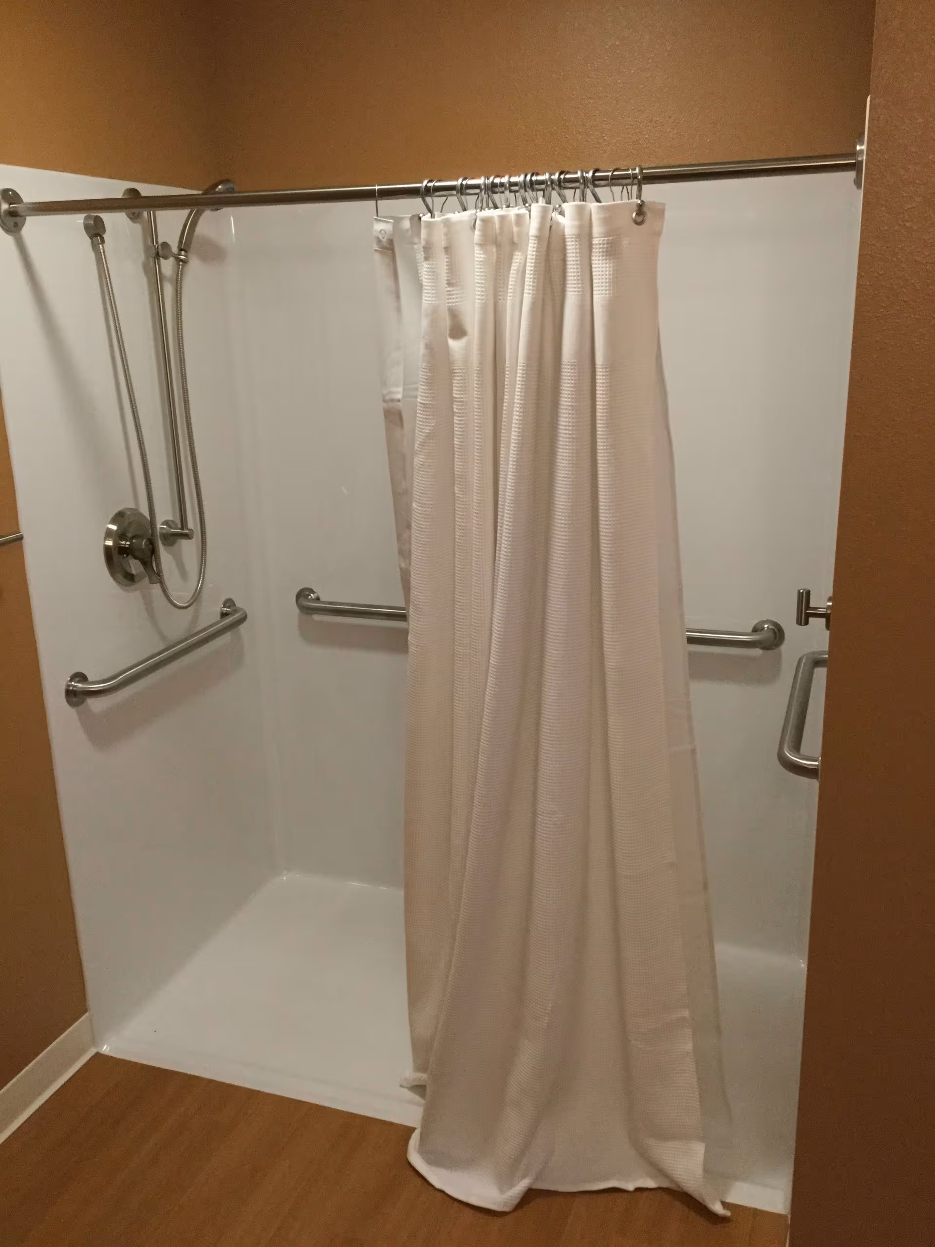 A walk-in shower with white walls and a white shower curtain partially drawn. The shower has a handheld showerhead and multiple stainless steel grab bars installed on the walls for support. The floor outside the shower is wood-toned.