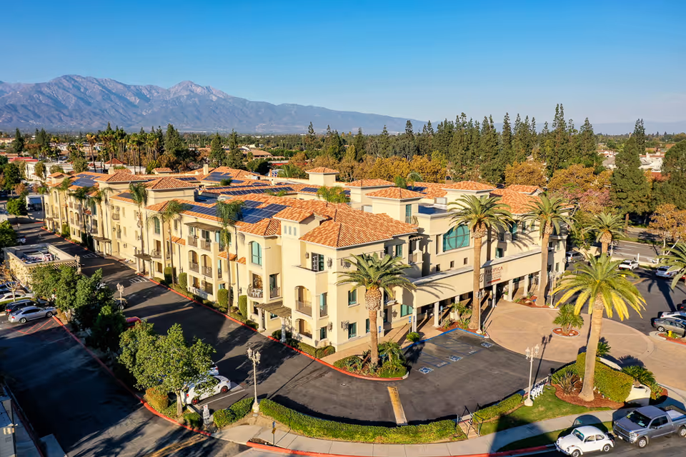 Aerial view of a Mediterranean-style senior living building with palm trees, parking lot, and mountains in the background.