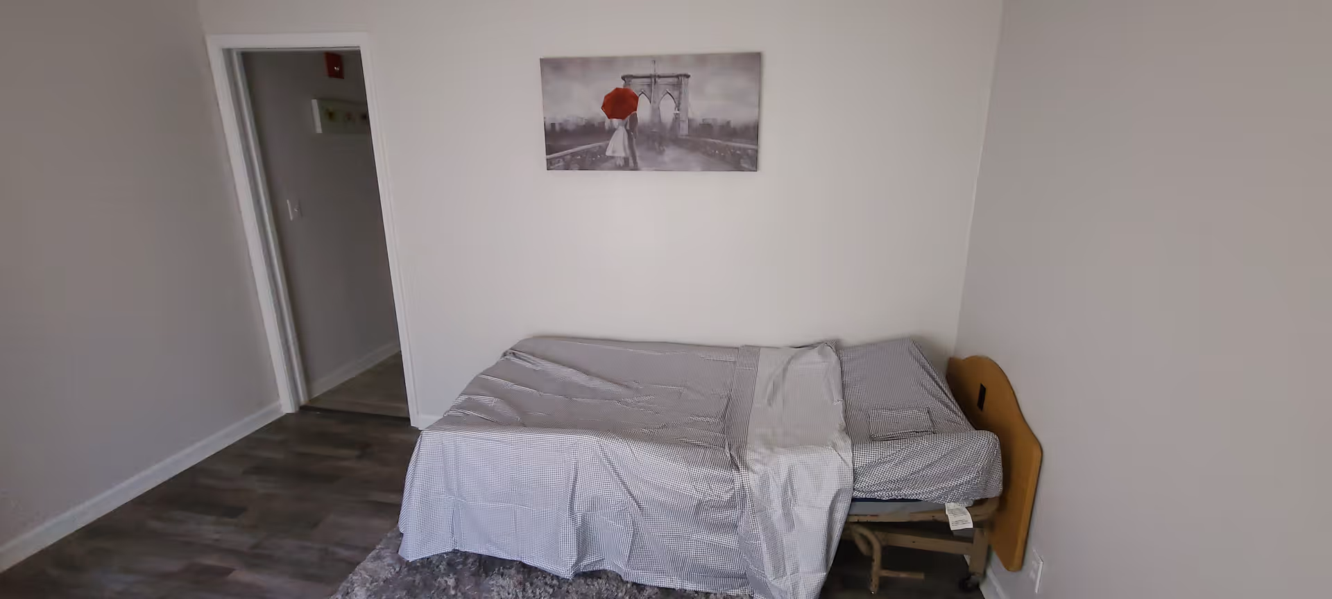 A small bedroom with a single bed against the right wall, covered with a gray sheet and a folded blanket. Above the bed hangs a black and white picture of a couple under a red umbrella on a bridge. The floor is wooden with a gray rug partially visible. There is an open doorway leading to another room on the left side.